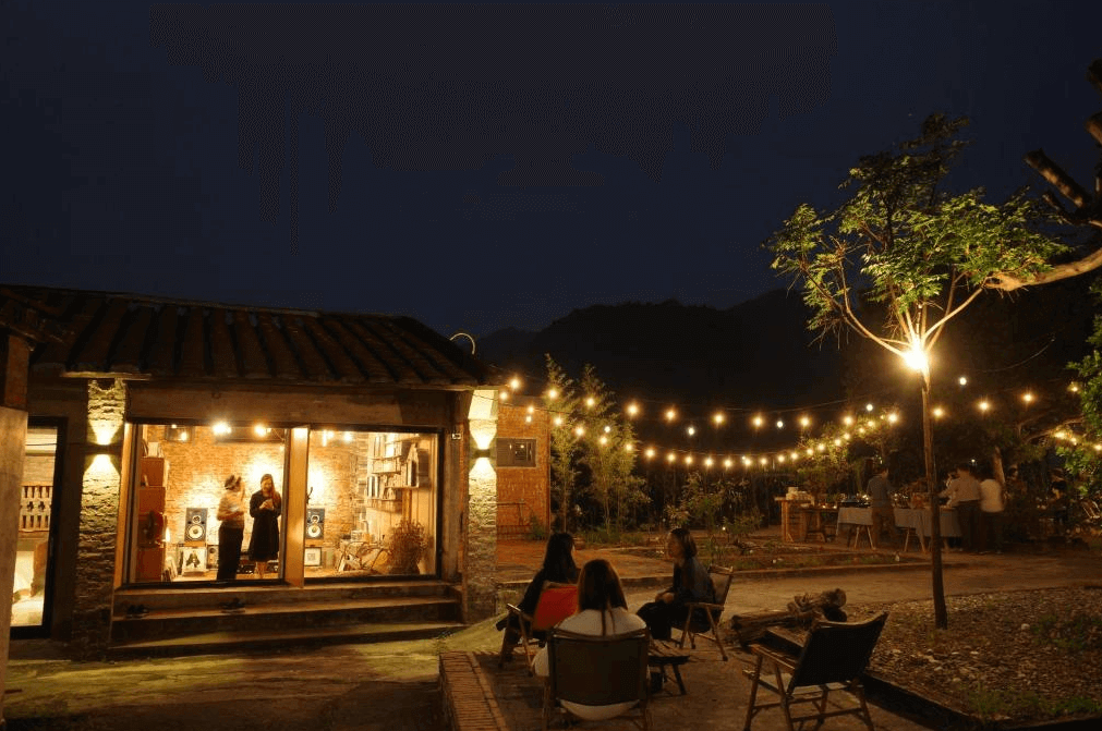 【美濃住宿】香蕉與黑膠三合院的聚會區，夜晚時燈光環繞，氣氛溫馨。人們圍坐在燈光下的院子裡，享受輕鬆的時光，背景是一間裝飾有許多黑膠唱片和音響設備的小屋，透過大窗戶可以看到屋內的音樂休閒區。