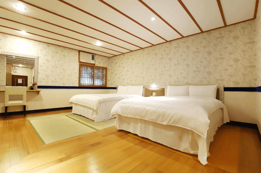 麗之湯溫泉會館雙人房：一間設有雙人床的客房，房內有兩張白色床單和枕頭，地板為木質地板，牆壁上有花卉圖案的壁紙，整體氛圍溫馨且舒適。房間內還有一個梳妝台和鏡子，設計簡約且實用