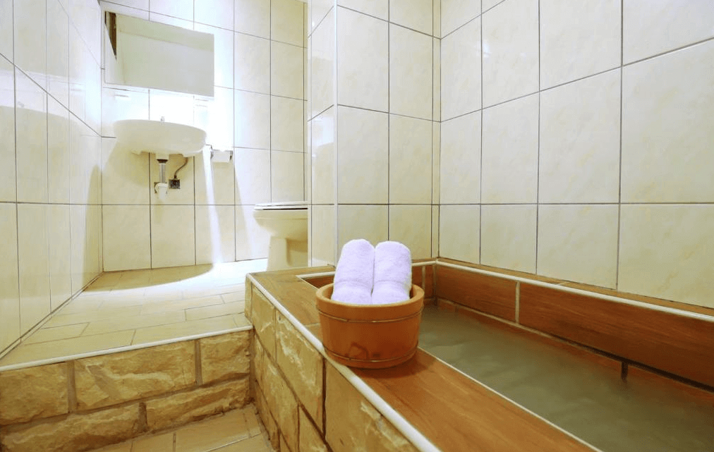 麗之湯溫泉會館私人浴室：一間設有木製浴缸的私人浴室，牆壁和地板為淺色瓷磚。浴室內擺放著毛巾和木桶，提供舒適的洗浴體驗，整體設計簡約且實用