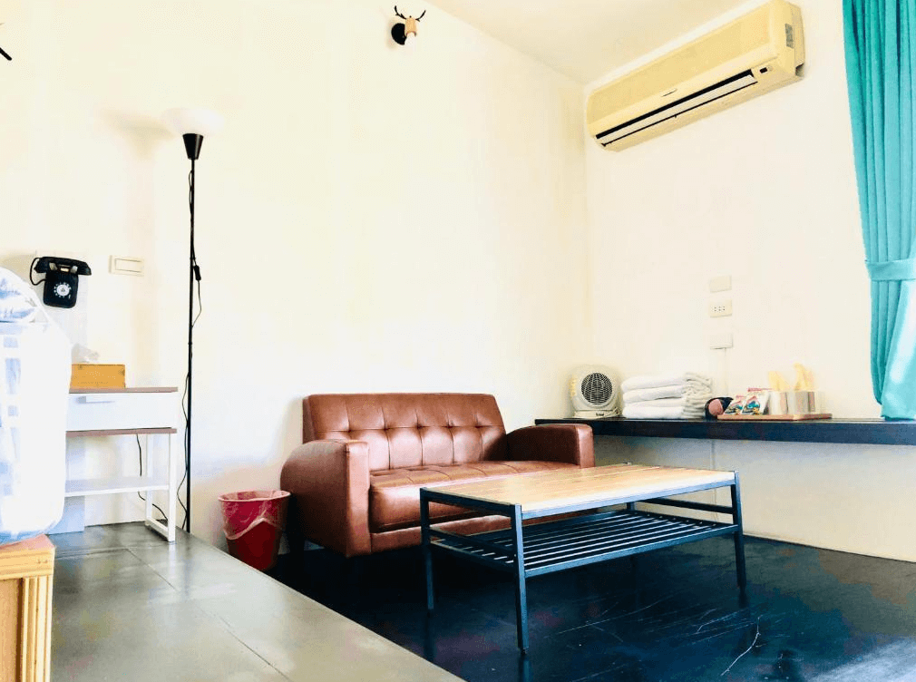 【九份寵物友善住宿】九居的溫馨客廳，內部設有一張棕色皮質沙發和一個木質茶几。牆上掛有一台老式電話和一些裝飾，房間角落擺放著落地燈，空調位於牆上，窗簾為藍綠色。