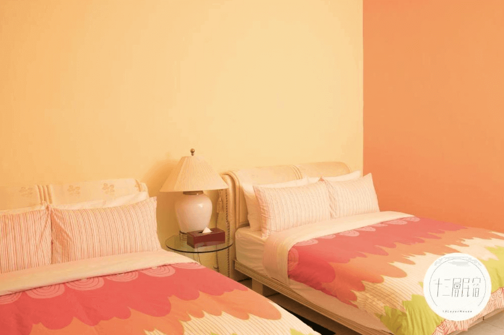 【九份寵物友善住宿】十三層民宿的雙床房，內部設有兩張舒適的大床，床上鋪有粉色和橙色相間的被褥。房間牆壁為柔和的黃色和橙色，配有一盞溫暖的床頭燈，營造出溫馨舒適的氛圍。