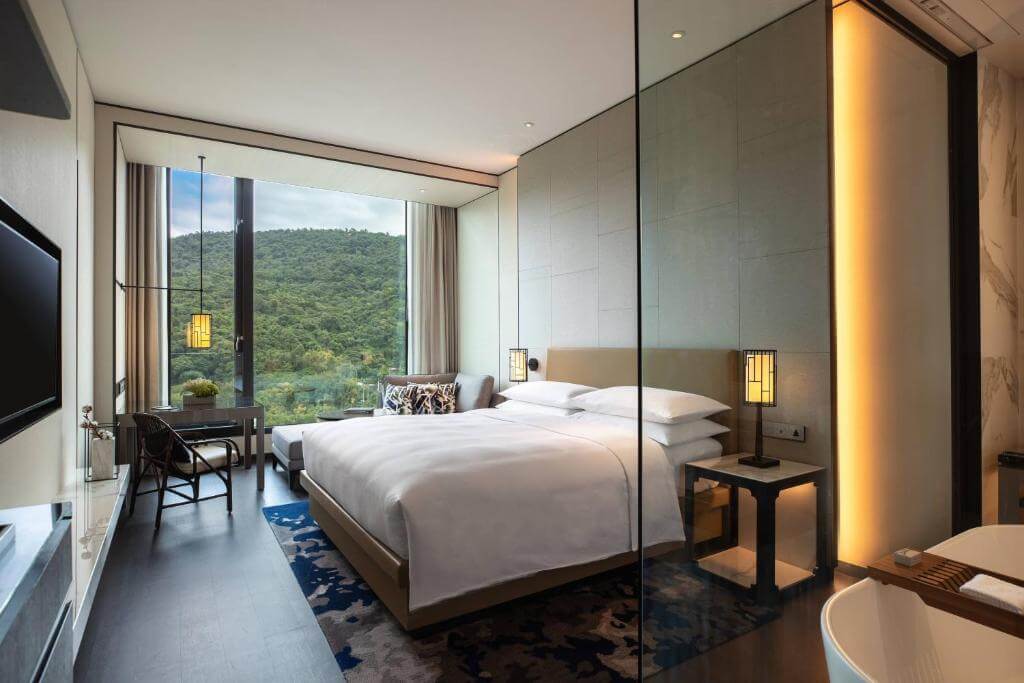 【台北親子飯店】台北士林萬麗酒店展示了一間裝飾現代的酒店客房。房間內有一張大床，床上鋪著白色的床單，兩側各有一個床頭燈。房間的一側是大落地窗，窗外可以看到郁郁蔥蔥的山景。窗前有一個小桌子和椅子，適合休息或工作。整個房間設計簡約大方，色調柔和，給人一種舒適和放鬆的感覺。