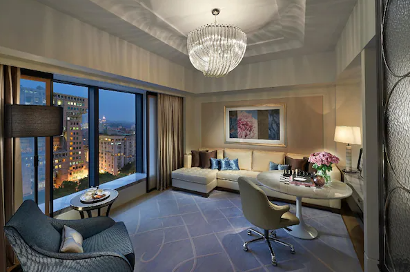 【台北親子飯店】台北文華東方酒店客廳 這張照片展示了酒店內一個寬敞且高雅的客廳。客廳內設有一組舒適的沙發和一個圓形的桌子，適合休閒和工作。窗外可以看到城市的夜景，天花板上的水晶吊燈和牆上的藝術畫作增添了房間的藝術氣息和高級感。