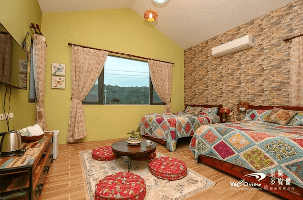 吾居吾素展示了一間溫馨的雙人房，房間內有兩張雙人床，床上鋪著彩色拼布床單。房間內還有一個小圓桌和幾個坐墊，牆面採用石頭裝飾，窗外可以看到山景