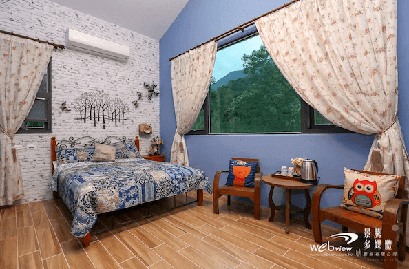 吾居吾素展示了一間帶有森林主題的客房，房間內設有一張雙人床和兩把木製椅子，床上鋪著藍色花紋的床單。窗外可以看到綠樹成蔭的風景，牆上掛有一幅樹林壁畫，營造出自然的氛圍