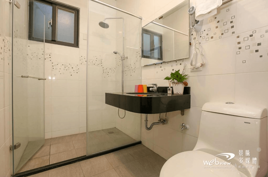 吾居吾素展示了一間現代化的浴室，浴室內設有玻璃淋浴間和一個黑色洗手台，台面上擺放著綠色植物和幾個彩色杯子。牆上還有一面鏡子，整體設計簡潔且實用