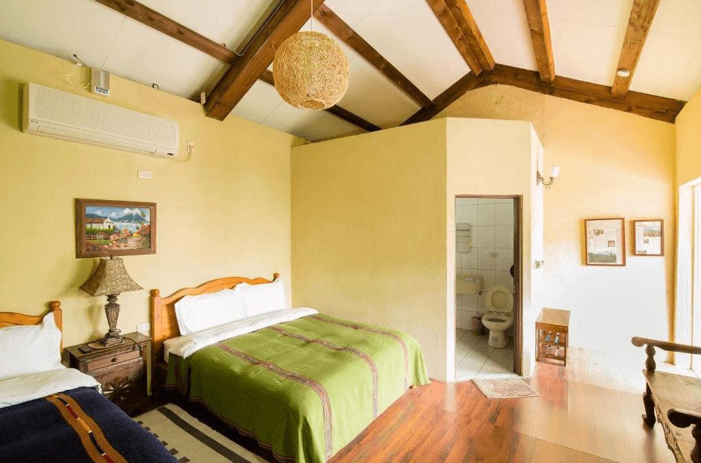 花蓮莫內花園民宿展示了一間溫馨的雙人房，房間內有兩張雙人床，床上鋪著綠色床單。牆面上掛著一幅風景畫，空調裝置安裝在牆上。房間內有木質天花板，設計簡約溫暖，右側有一個小衛生間