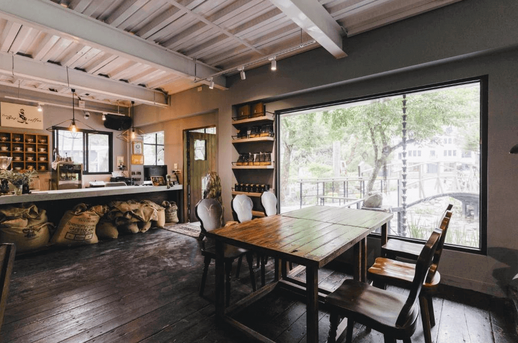 花蓮莫內花園民宿展示了一間舒適的咖啡廳，內部裝飾以木質為主，桌椅擺放整齊。大窗戶讓自然光線充足進入，窗外可以看到綠意盎然的庭院。櫃檯後方擺放著咖啡豆袋和各種咖啡器具，營造出溫馨的氛圍