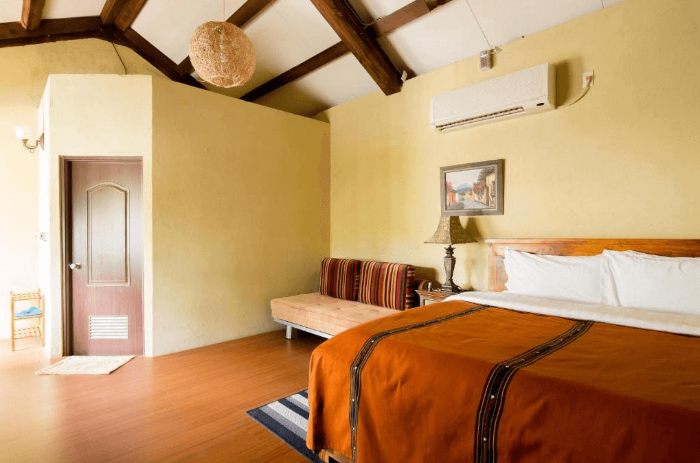 花蓮莫內花園民宿展示了一間寬敞的客房，房間內有一張大床，床上鋪著橙色床單。旁邊有一個小沙發和一盞溫暖的燈。天花板上有木質橫樑，設計風格自然簡約，房間一角有一個小衛生間