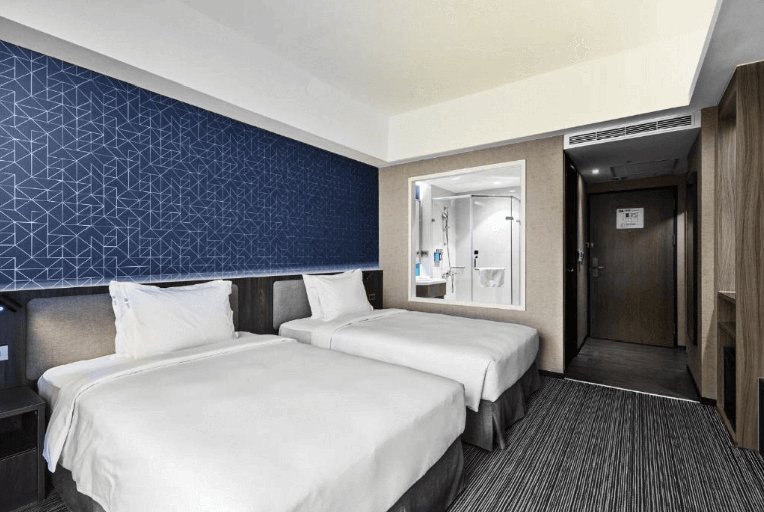 高雄愛河智選假日酒店的雙床房，房間內裝潢現代且簡約，牆面設有幾何圖案背景牆，房間內設有兩張單人床，床鋪整潔舒適，提供優質的住宿環境。浴室部分採用透明玻璃設計，讓房間顯得更為寬敞明亮。