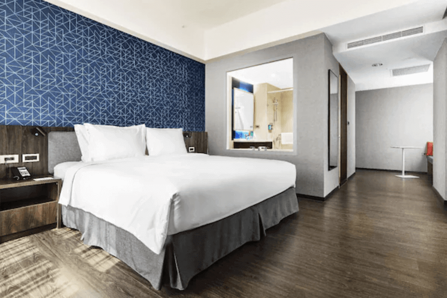 高雄愛河智選假日酒店的雙人房，房間內設有一張大床，床鋪整潔舒適。背景牆同樣採用幾何圖案設計，整體風格現代時尚。房間內部寬敞明亮，提供舒適的住宿環境。浴室部分亦採用透明玻璃設計，增加空間感