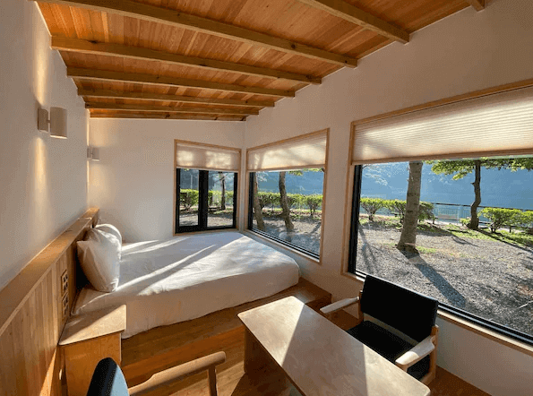 黑屋CAMP展示了一間現代風格的客房，房間內設有一張雙人床，床邊的窗戶外可以看到湖景和樹木。房間內有大量自然光照射進來，營造出溫暖的氛圍