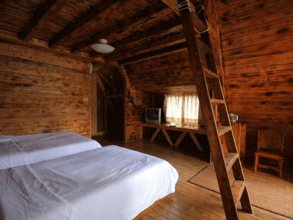 黑屋CAMP展示了一間木屋風格的客房，房間內設有兩張雙人床，並有一個木製樓梯通往上層。房間內有大量木質裝飾，營造出溫暖舒適的氛圍