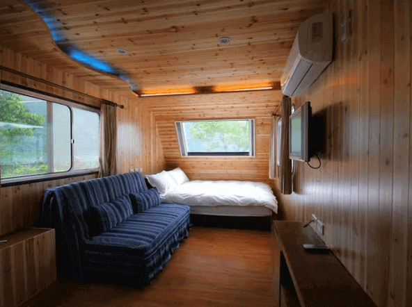 展示了一間木質內裝的客房，房間內設有一張藍色條紋的沙發和一張舒適的雙人床，床尾有一台平板電視，窗戶外可以看到部分戶外景色