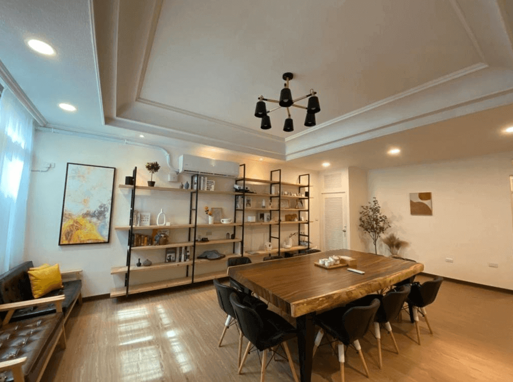 千柏旅居展示了一個現代風格的公共用餐區。房間內有一張大木質餐桌和多張椅子,周圍的書架上擺放著書籍和裝飾品。牆上掛有現代藝術畫作,整體感覺溫馨且舒適,適合家庭或朋友聚會。