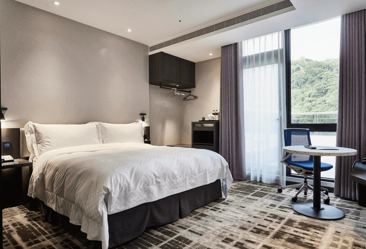 天閣酒店-台北士林館展示了一間現代風格的酒店客房。房間內有一張大床，床上鋪著白色床單，顯得非常乾淨舒適。牆壁是淡雅的灰色，房間內有一張小桌子和椅子，適合工作或休息。窗戶掛著紫色的窗簾，窗外可以看到綠意盎然的景色，讓房間感覺明亮和開闊。