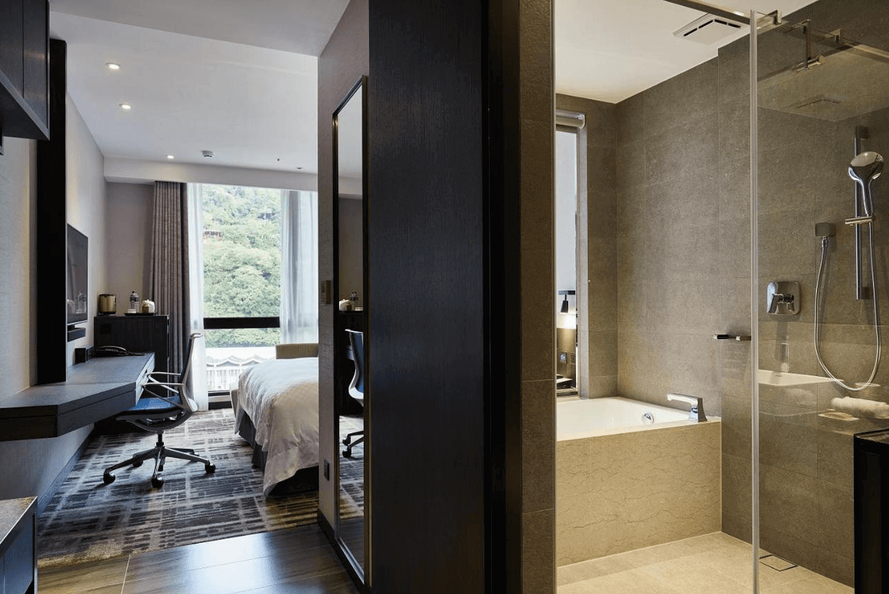 天閣酒店-台北士林館展示了一間現代風格的酒店客房及其浴室。房間內有一張大床，床上鋪著白色床單，顯得非常乾淨舒適。房間內部設計簡約，牆壁是淡雅的灰色。浴室內有一個獨立的浴缸和淋浴區，浴室與房間之間有一個玻璃門。整體設計給人一種舒適和現代的感覺。