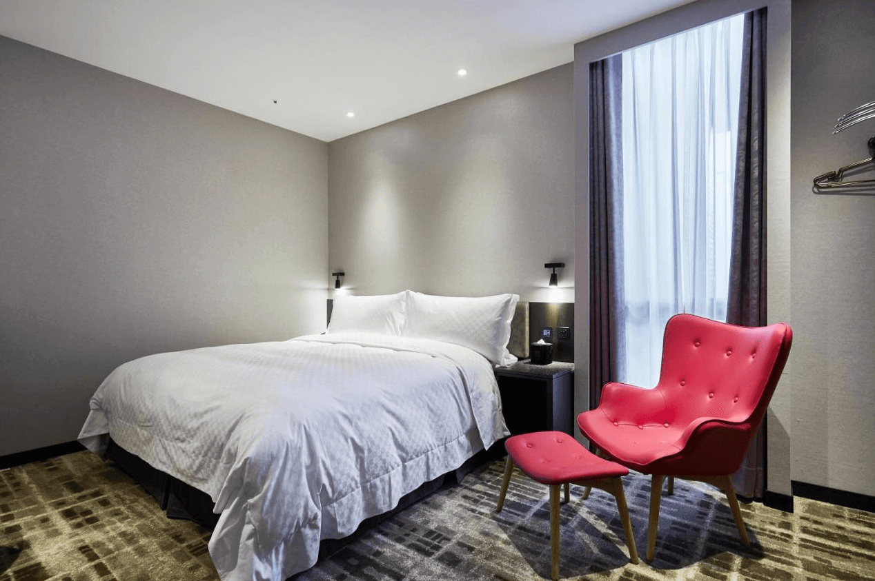 天閣酒店-台北士林館展示了一間現代風格的酒店客房。房間內有一張大床，床上鋪著白色床單，顯得非常乾淨舒適。房間內有一張鮮紅色的休閒椅和腳凳，為房間增添了一抹亮色。窗戶掛著紫色的窗簾，整體設計簡約而溫馨。