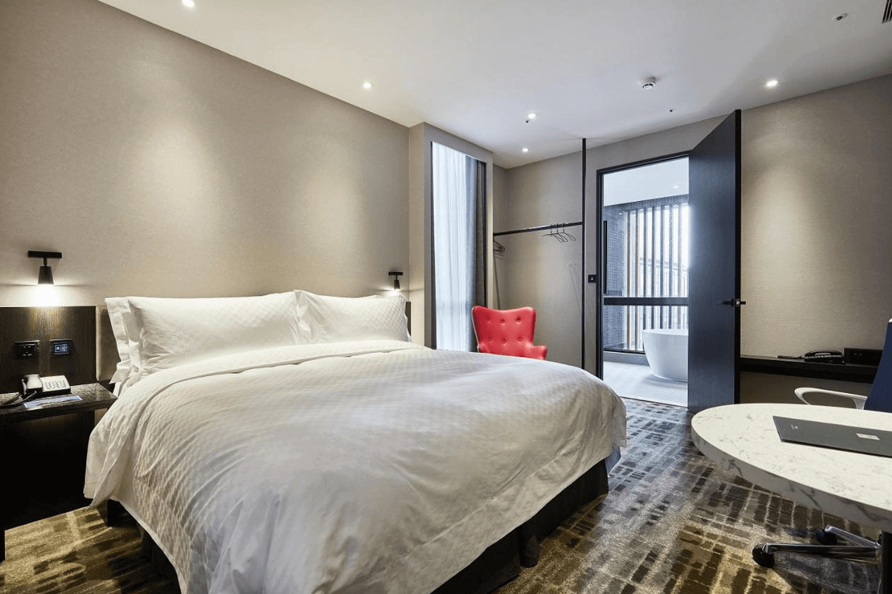 天閣酒店-台北士林館展示了一間現代風格的酒店客房。房間內有一張大床，床上鋪著白色床單，顯得非常乾淨舒適。房間內還有一張小桌子和椅子，適合工作或休息。窗戶掛著紫色的窗簾，窗外可以看到外面的景色。整體設計簡約大方，給人一種舒適的感覺。