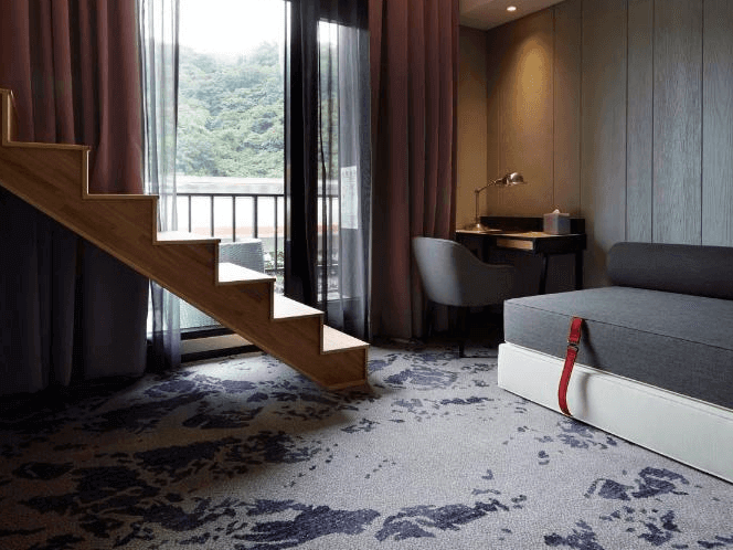 天雲旅棧-台北基河展示了一間現代風格的酒店客房，客房內有一個小型的樓梯，通往上層空間。房間內部採用紫色和灰色的色調，營造出舒適和溫馨的氛圍。房間內有一張單人沙發和一張小桌子，適合休息或工作。大窗戶讓自然光充滿整個房間，窗外可以看到郁郁蔥蔥的山景。