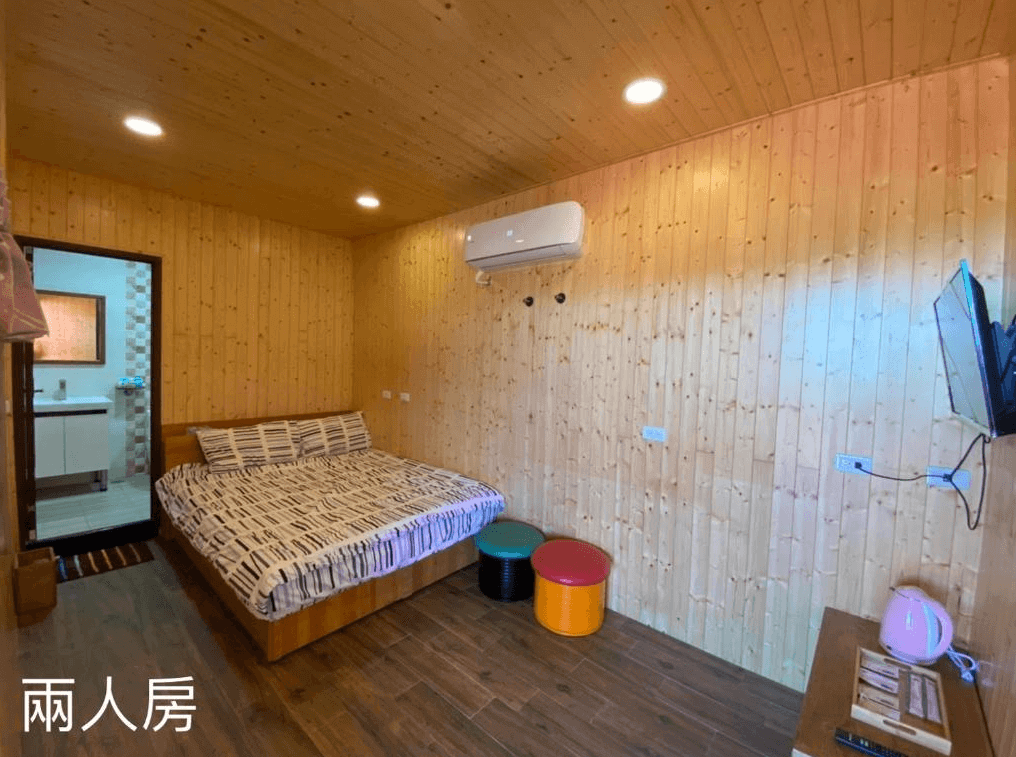 鎮安粉紅豹的雙人房內部,房間同樣使用木質牆壁和天花板設計,營造出溫暖的氛圍。房間內有一張大床、一台掛牆電視和空調,浴室也在房內,設計簡約實用。