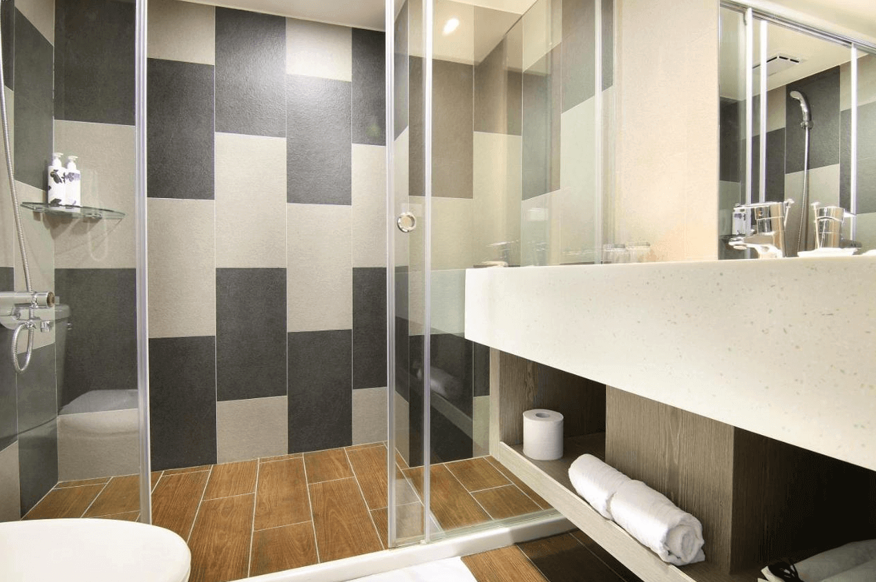 陽光士林珮柏旅館展示了一間現代風格的浴室。浴室內有一個寬敞的淋浴區，淋浴區的牆壁上採用灰色和米色相間的方格瓷磚，地板是木紋瓷磚。洗手台旁邊有一面大鏡子，洗手台下方有開放式的儲物空間，用於放置毛巾和衛生紙。整體設計簡約實用，顯得非常乾淨和現代。