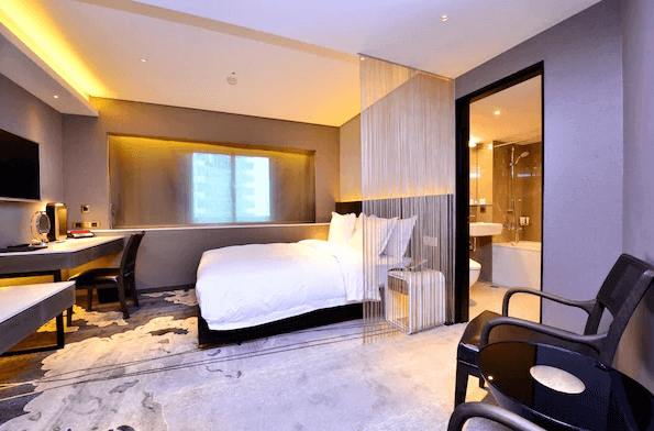 【台北小巨蛋住宿】兄弟大飯店的客房，內部設計簡約，具有一張大床，床頭牆上設有柔和的黃光照明，增添了房間的溫暖感。房間中間有一道透明的玻璃牆將臥室與浴室隔開，玻璃上懸掛著一道輕薄的窗簾，提供隱私保護。浴室內部採用現代設計，整體氛圍舒適且乾淨。