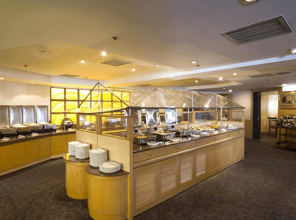 "台北小巨蛋住宿"兄弟大飯店的自助餐廳的圖片，餐廳內部空間寬敞明亮，設有長形自助餐檯，擺放著各種餐具和食物。餐檯上方採用透明玻璃頂棚設計，讓顧客可以清楚地看到食物的擺放情況，整體感覺乾淨整潔，充滿現代感。