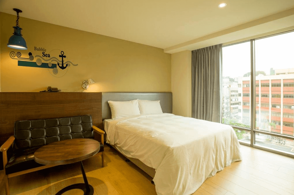 享住旅店房間內部的照片，展示了一張雙人床，牆上有海洋主題的壁畫，旁邊放置了一張小圓桌和黑色沙發椅。窗外光線明亮，整個房間氛圍溫暖舒適，設計簡潔。