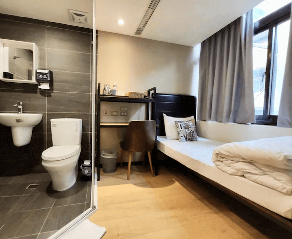 享住旅店一間小型客房的內部照片，房間內有一張單人床，旁邊設有小型書桌，整體空間緊湊而實用。房間內的浴室配有現代化的淋浴設備，採用深色瓷磚裝飾。