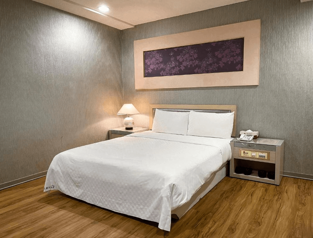 北都大飯店一間單人房的內部照片，房間內有一張雙人床，牆壁上方懸掛著一幅花卉畫作。床頭兩側有床頭櫃，一盞桌燈發出柔和的光線，地板是溫暖的木質材質。