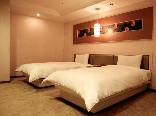 北都大飯店雙床房的內部照片，房間內有兩張雙人床，牆壁上方裝飾著一幅花卉畫作。床頭上方有吊燈照明，房間設計簡約且溫馨。