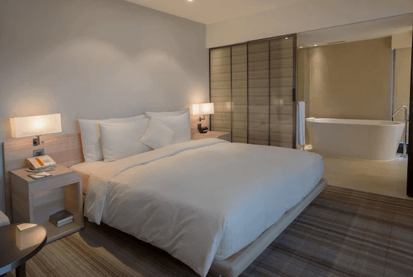 【松山機場住宿】和逸飯店-台北民生館的酒店客房，房內配有一張大床，床頭背景牆顏色柔和，營造出溫馨的氛圍。房間內設有木質床頭櫃和柔和的燈光，提供舒適的休息環境。房間與浴室之間由半透明的玻璃門隔開，浴室內可見到一個獨立式浴缸，設計典雅。