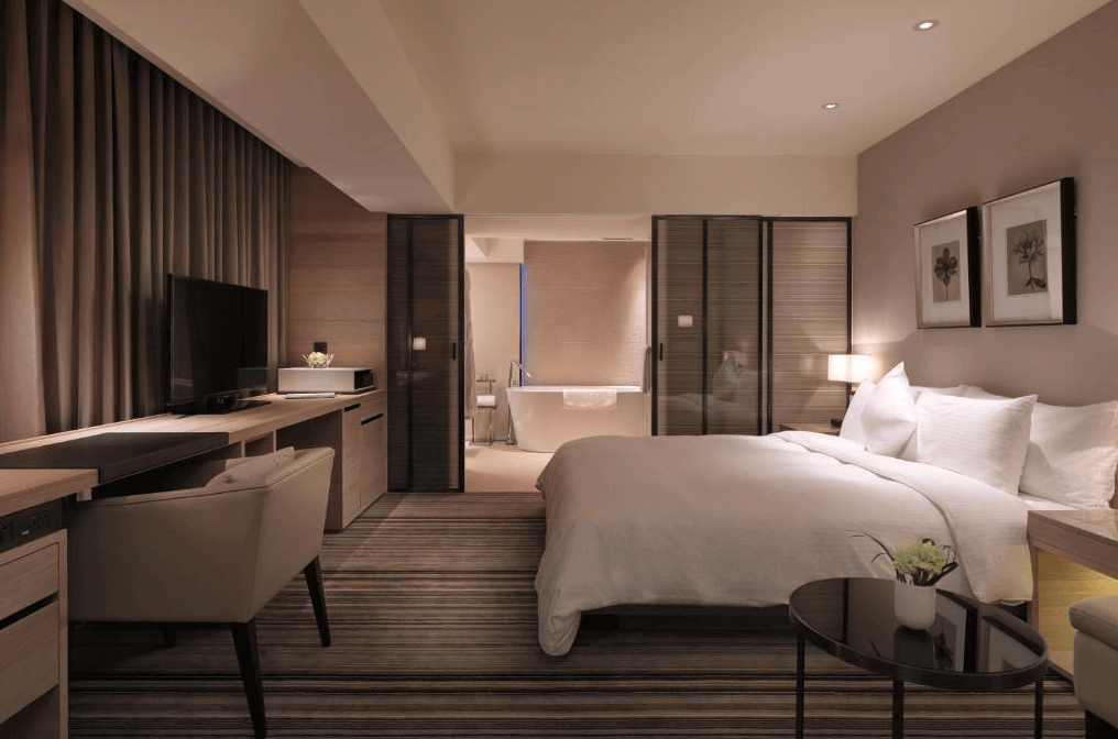 【松山機場住宿】和逸飯店-台北民生館的圖片，設計簡約而不失奢華。房內配有一張大床，床邊設有柔和的燈光和現代化家具。房間內還設有一個辦公區，並且可以看到玻璃門後的浴室，浴室內配有一個獨立式浴缸，整體設計強調舒適與放鬆。