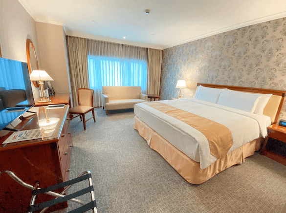 基隆長榮桂冠酒店的房間內部的照片，顯示一張大雙人床，床頭有兩盞床頭燈，牆壁上有花紋壁紙，窗邊放置了一張沙發和兩把椅子。房間佈置典雅，窗簾遮擋部分自然光線。