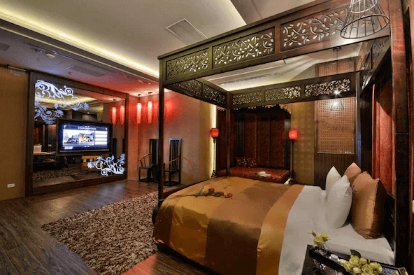 【台中情趣汽車旅館】夏都汽車旅館房間設計融合了中式古典與現代風格，擁有精美雕花的四柱床，床上鋪著金色和棕色的被褥。房間內的燈光設計溫馨，牆面和地毯的細節增添了奢華感。電視區和床頭燈具的設計更是凸顯了高雅的氛圍。