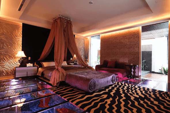 【台中情趣汽車旅館】夏都汽車旅館房間設計奢華且充滿現代感，地板上設有玻璃和燈光裝飾，營造出夢幻般的氛圍。床上鋪著柔軟的紫色毛毯，搭配金色和棕色的牆面裝飾，讓人感到既舒適又高雅。整體設計讓房間充滿浪漫和奢華感。