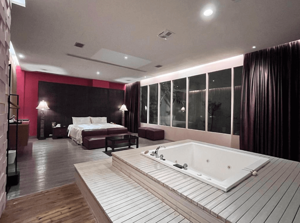 【台中情趣汽車旅館】天月人文休閒汽車旅館房間寬敞明亮，擁有豪華的大床和按摩浴缸。床頭牆面以深色為主，搭配紅色的背景牆，增添了一份浪漫的氛圍。大面積的窗戶讓自然光線充滿整個房間，提供舒適的視覺享受。