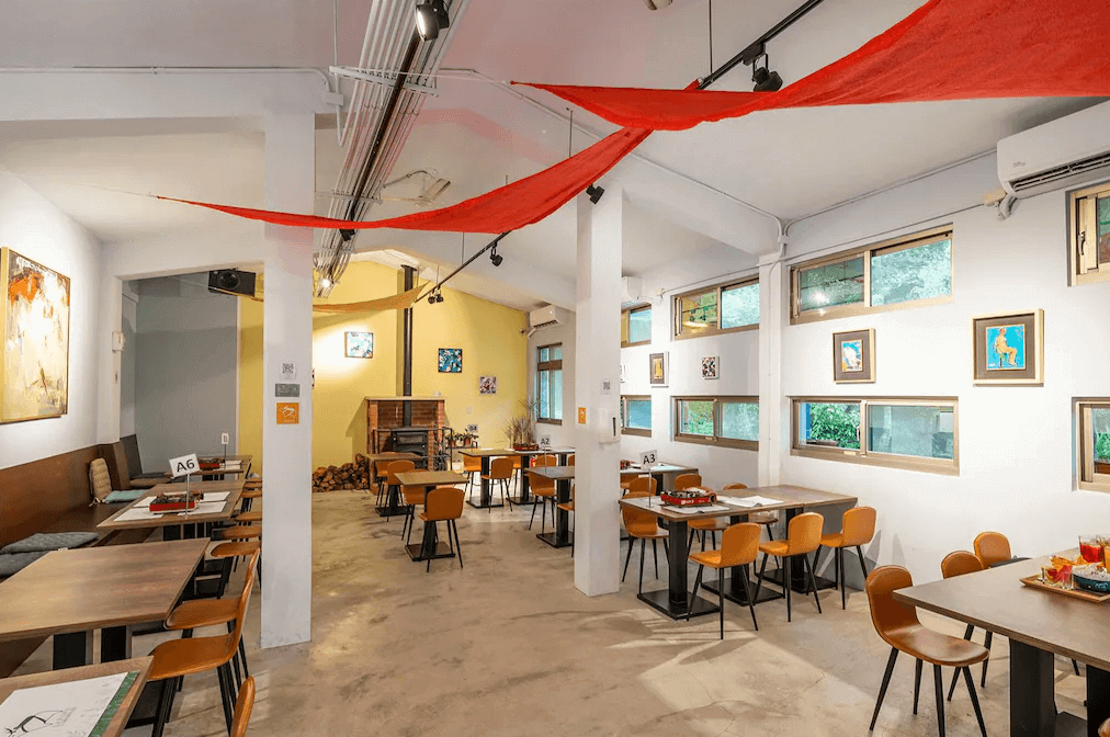 拉波波村飯店的餐廳的內部環境，白色牆壁上掛著多幅裝飾畫，紅色的裝飾布掛在天花板上。餐廳內擺放著多張木桌和木椅，牆邊有一個壁爐，整體氛圍溫馨而有藝術感。