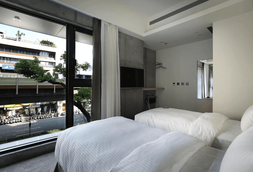 【松山機場住宿】新驛旅店-復興北路店的雙床客房，房間內部設計簡約，配有大面積的落地窗，可以看到外面的城市景觀。窗戶旁邊有兩張白色床鋪，床單整潔，光線充足。牆壁採用灰色混凝土質感，增添了一些工業風格。