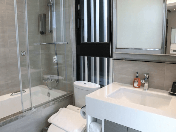 【松山機場住宿】新驛旅店-復興北路店的浴室，內部設有淋浴區和浴缸。牆壁和地板都以淺灰色瓷磚裝飾，顯得非常乾淨。洗手台設計簡潔實用，旁邊放有毛巾和基本的衛浴用品，窗戶提供了自然光線，讓空間感更為開闊。