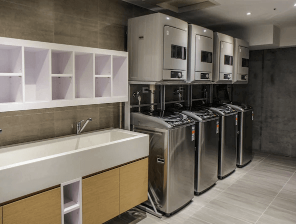 【松山機場住宿】新驛旅店-復興北路店的自助洗衣房，設有多台洗衣機和烘乾機，整齊排列。洗衣房內部空間寬敞，配有一個大水槽，牆面和地板都採用灰色瓷磚裝飾，整體感覺非常整潔和實用。