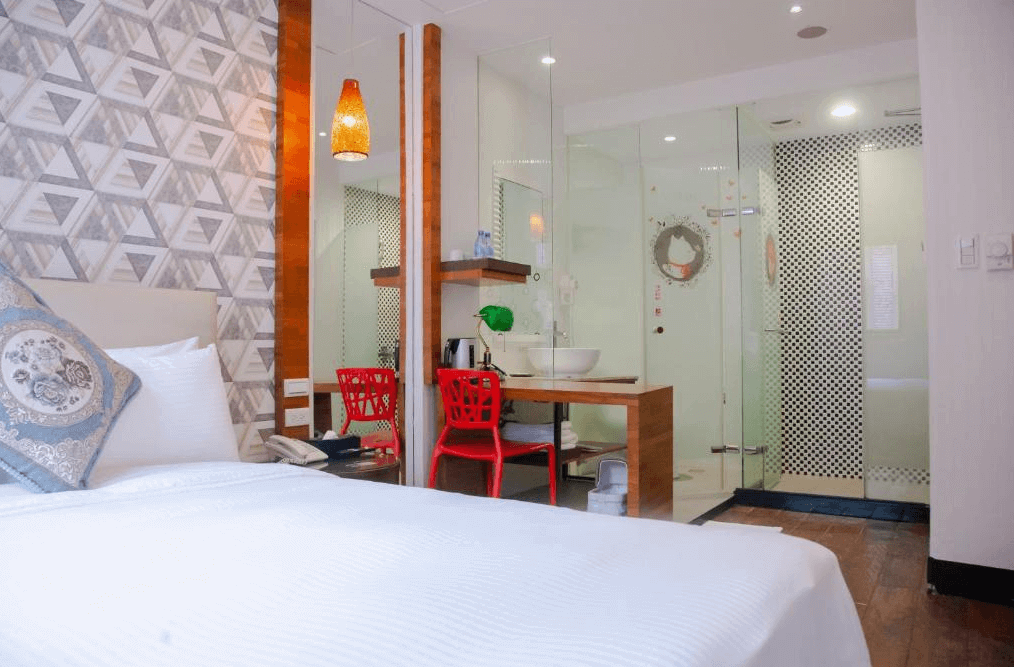 房間內部的照片，顯示了一張雙人床，床頭背景牆使用了幾何圖形壁紙，房間內設有一張紅色椅子和木質桌子，桌子上方設有一個白色洗手盆，旁邊是玻璃隔間的浴室。設計現代且色彩鮮明。