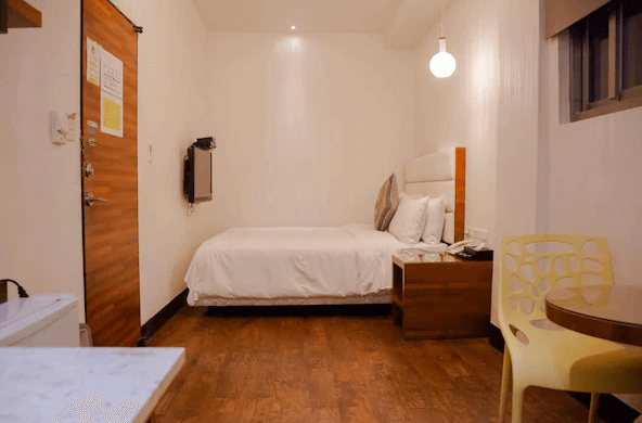 一間小型單人房的內部照片，房間內有一張單人床，牆壁採用簡單的白色設計，房內設有一張小桌子和黃色椅子，地板是深色木質材料，空間佈置緊湊而溫馨。