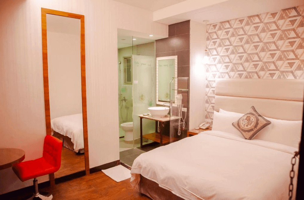 一間雙人房的照片，房內設有一張雙人床，床頭背景牆為幾何圖形設計，床旁邊是一個帶有玻璃隔間的浴室，房間內還有一張紅色椅子和小圓桌，氛圍溫馨且充滿現代感。