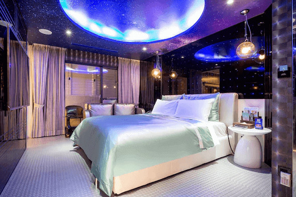 【台中情趣汽車旅館】極光情境旅館房間設計充滿未來感和科技感，天花板上的藍色燈光效果營造出星空般的氛圍。房間內擺設著一張舒適的大床，搭配銀色和白色的家具，整體感覺現代且時尚。大窗戶讓自然光線充盈房間，提供明亮的視覺享受。