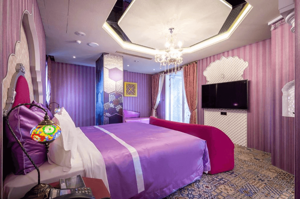 【台中情趣汽車旅館】極光情境旅館房間設計充滿異國情調，牆面和家具以紫色為主，營造出浪漫的氛圍。床頭有華麗的吊燈和色彩繽紛的燈具，增加了房間的視覺效果。整體設計富有藝術感且充滿魅力。