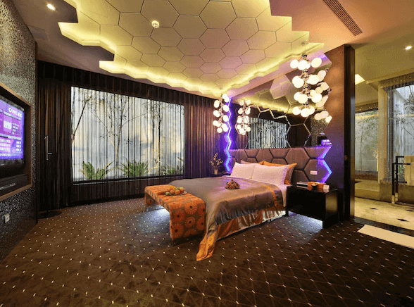 【台中情趣汽車旅館】涵館汽車旅館的房間設計充滿現代感和科技感，擁有一張大床，床頭裝飾著六邊形圖案的鏡面和燈具。天花板和床頭牆面上的燈光設計巧妙，營造出時尚的氛圍。大窗戶讓自然光線充盈房間，提供舒適的視覺體驗。