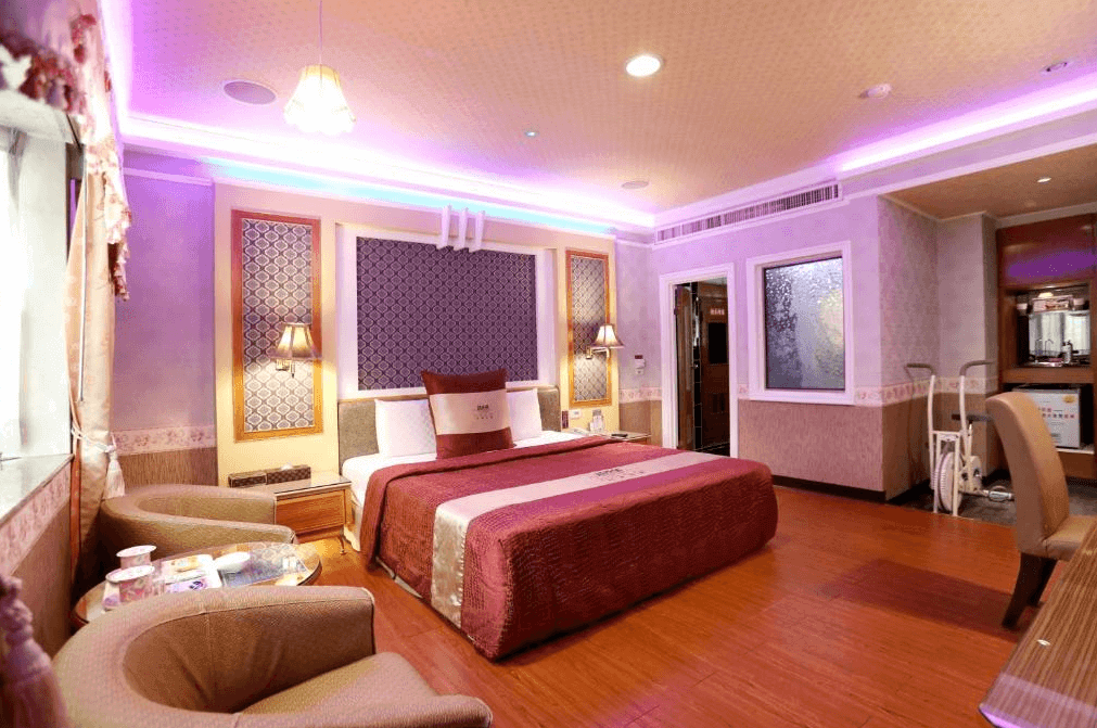 【新營汽車旅館】温莎堡汽車旅館新營館的房間裝飾華麗，牆面和天花板都有淡紫色燈光環繞，中央擺放著一張大床，床上鋪有紫紅色的床單和枕頭，房內設有兩張舒適的座椅以及窗邊的茶几。