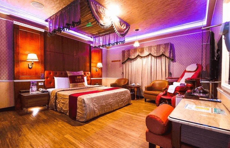 【新營汽車旅館】温莎堡汽車旅館新營館的房間內充滿浪漫氛圍，紫色燈光點綴著整個空間，中央的大床鋪有米色和紅色的床上用品，房內設有紅色按摩椅和兩張舒適的座椅，天花板懸掛著帷幔裝飾，增添了華麗的感覺。